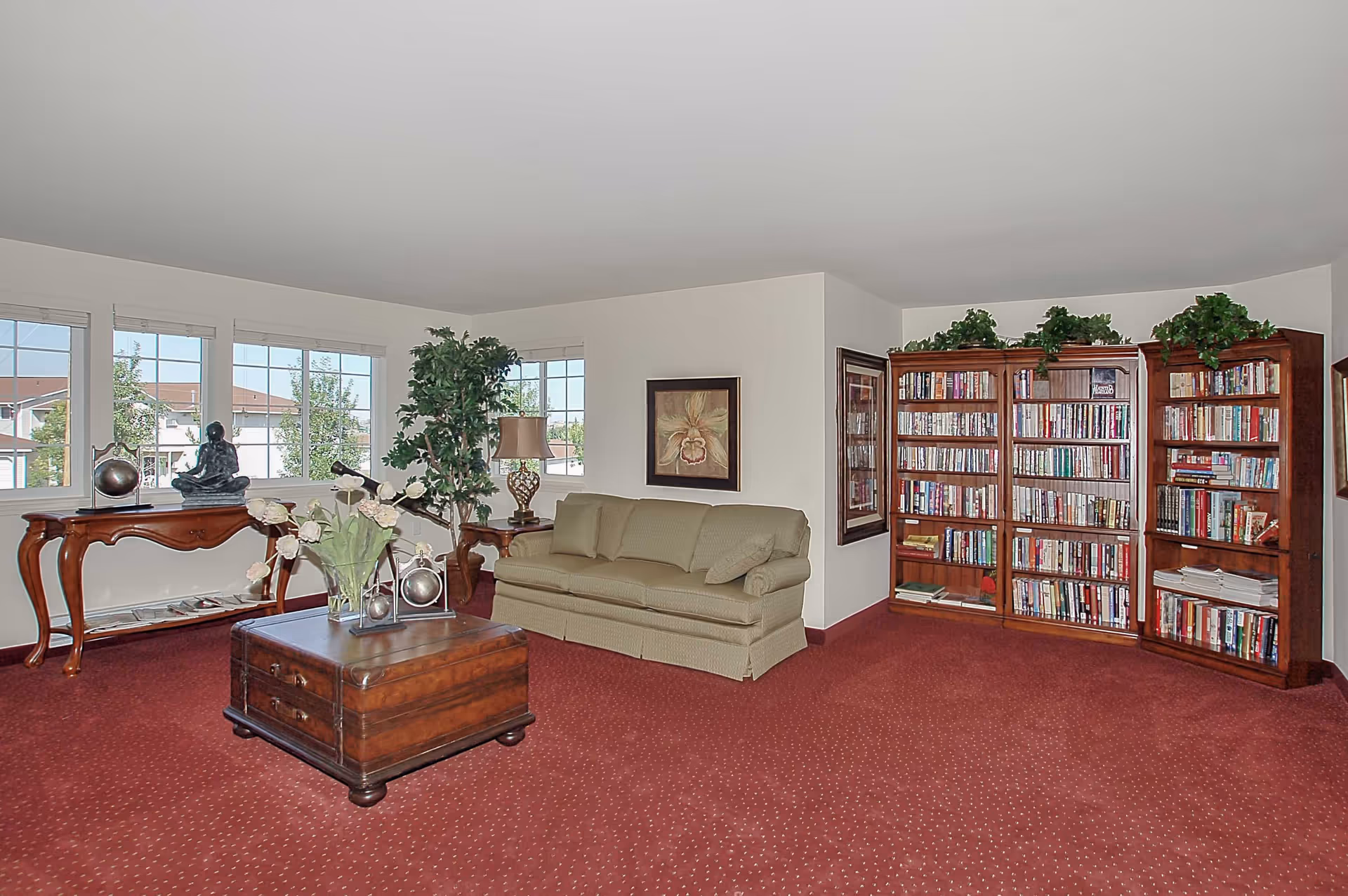 Spacious living room with a green sofa, wooden coffee table and side tables, large bookcases, decorative plants, and multiple windows over red carpeting.