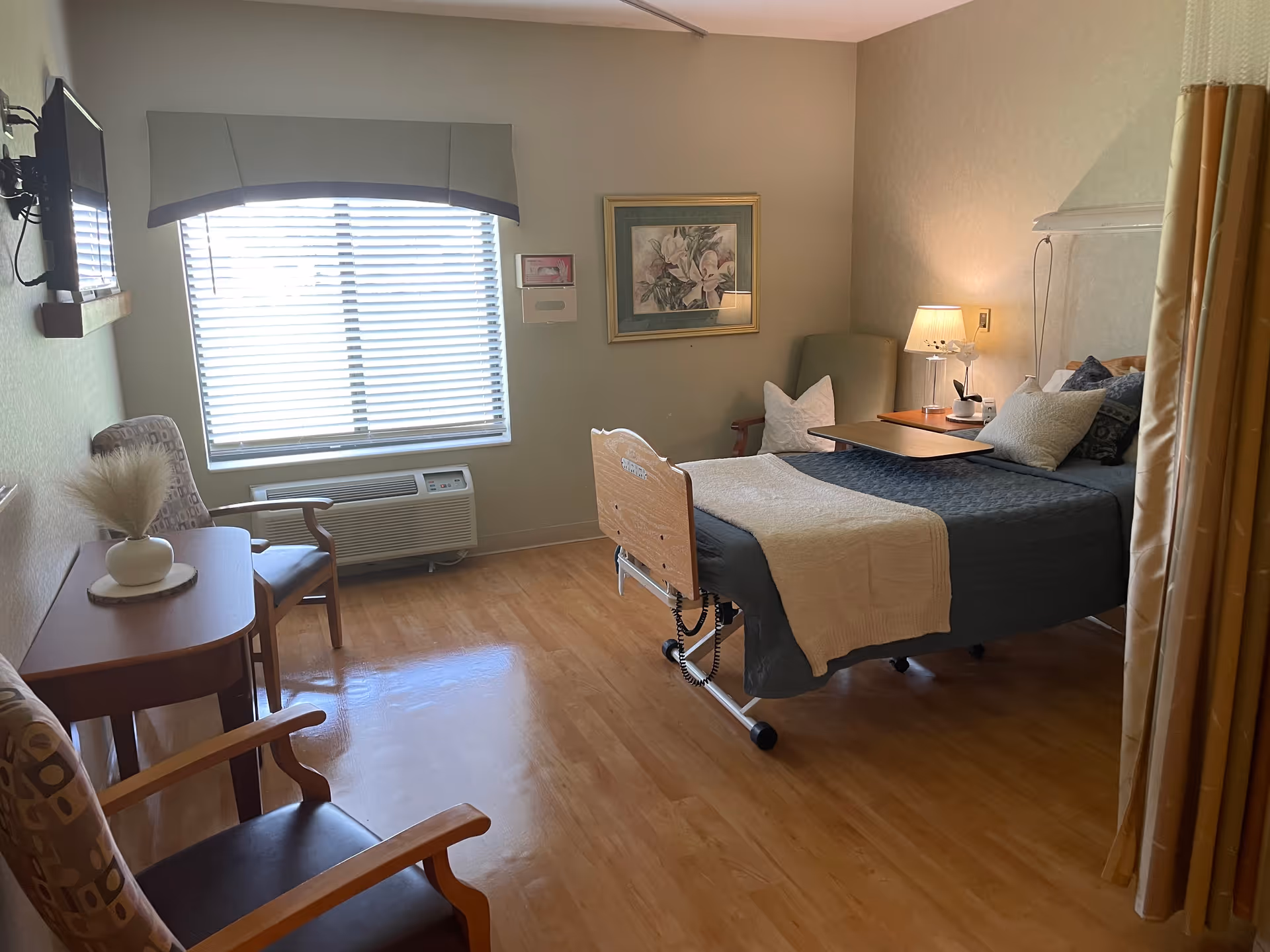 A senior care facility bedroom with a hospital-style bed covered with a dark blue blanket and a beige throw. There is a wooden bedside table with a lamp and a chair with a pillow next to the bed. Two wooden chairs and a small table with a decorative vase are near a window with blinds and a valance. The floor is wooden, and there is a framed floral picture on the wall.