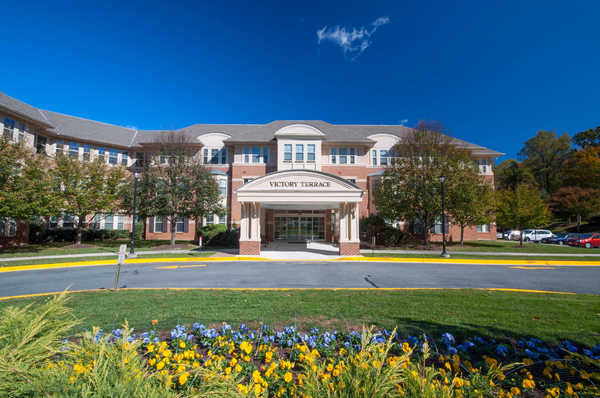 Front exterior view of Victory Terrace, a three-story senior living facility with a covered entrance, surrounded by trees and a flower bed with yellow and purple flowers under a clear blue sky.