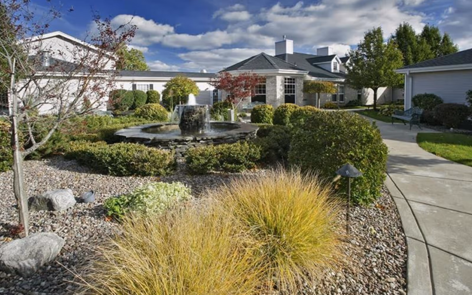 Outdoor garden area at Tennyson Court featuring a stone water fountain surrounded by neatly trimmed bushes, ornamental grasses, and trees under a partly cloudy sky. A concrete pathway curves through the landscaped garden with benches along the side.