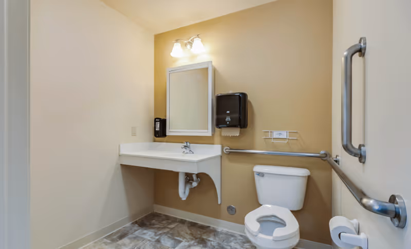 A clean and accessible bathroom with beige walls and tiled floor. The room features a white toilet with grab bars on both sides, a white wall-mounted sink with exposed plumbing, a mirror above the sink, a wall-mounted soap dispenser, and a paper towel dispenser. The lighting is provided by a double-bulb fixture above the mirror.