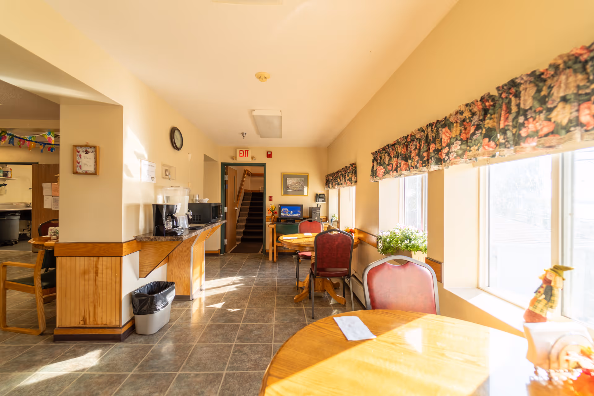 A bright dining area with round wooden tables and red cushioned chairs next to large windows with floral valances. A counter along one wall holds a coffee maker and microwave, with a trash bin underneath. The room has tiled flooring and cream-colored walls, with a clock and framed picture on the walls. An exit door and staircase are visible at the far end.