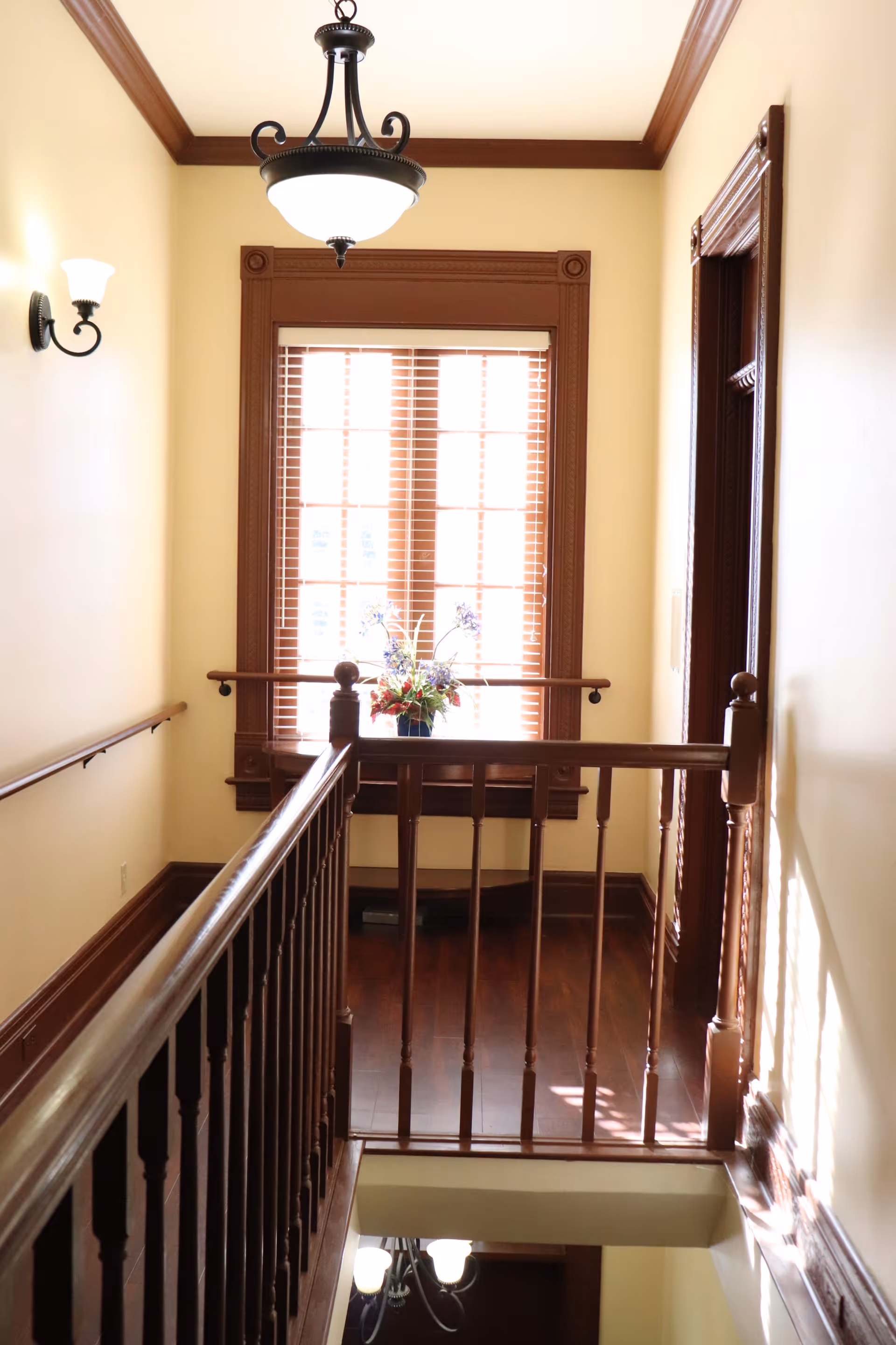 Interior view of a hallway with wooden railings and trim, a window with wooden blinds, a flower arrangement on the windowsill, and a ceiling light fixture.