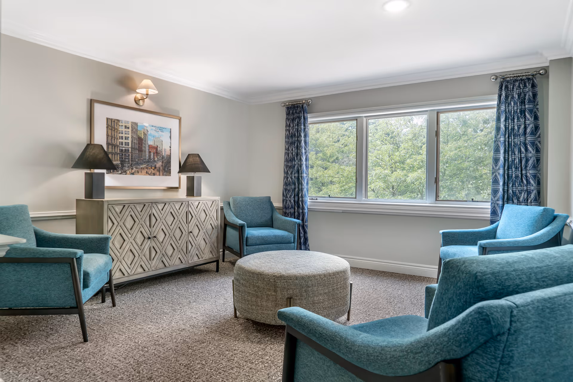 A bright living room with four teal armchairs arranged around a round gray ottoman. There is a decorative cabinet with two black lamps and a framed cityscape artwork above it. Large windows with blue patterned curtains let in natural light and show green trees outside.