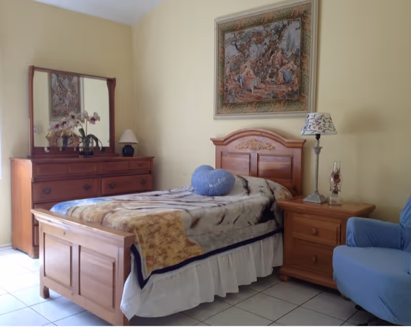 A cozy bedroom with a wooden bed featuring a patterned blanket and a blue heart-shaped pillow. To the right of the bed is a wooden nightstand with a decorative lamp and a glass ornament. On the left side of the room is a wooden dresser with a large mirror, adorned with a vase of flowers and a small lamp. A framed tapestry hangs on the pale yellow wall above the bed. A blue armchair is partially visible on the right side of the image.
