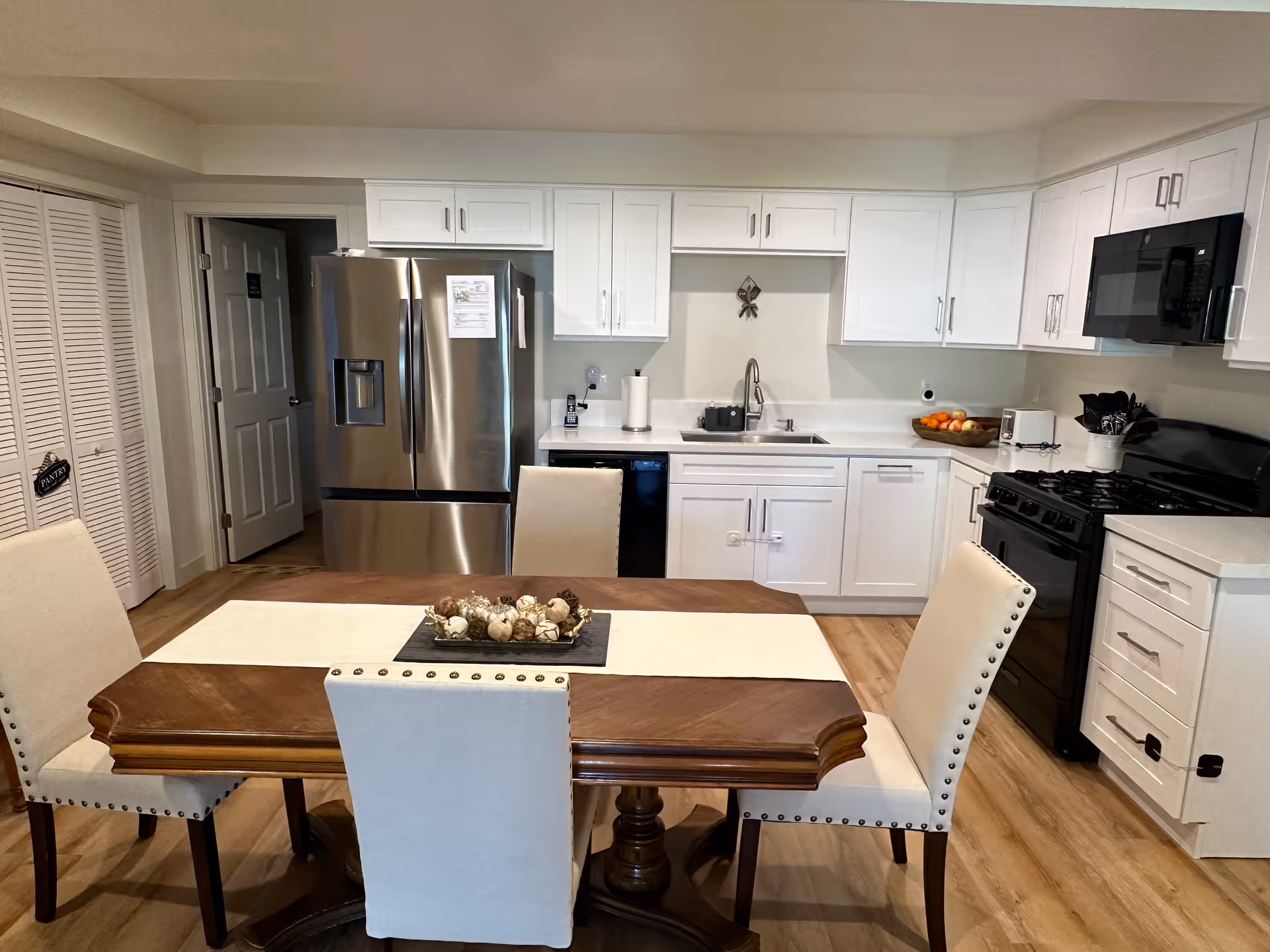 A modern kitchen and dining area with white cabinets, a stainless steel refrigerator, a black stove and microwave, and a wooden dining table with four upholstered chairs. The kitchen has a sink, countertop appliances, and a bowl of fruit on the counter.