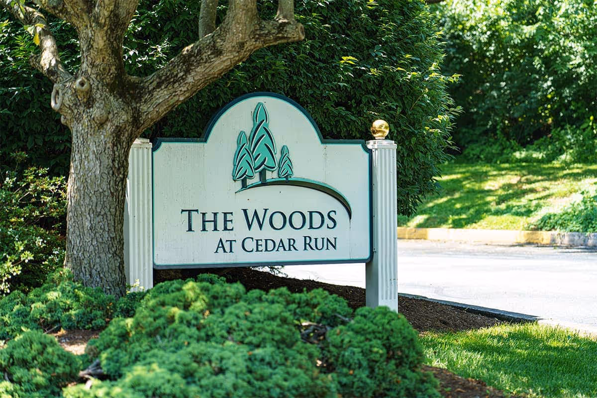 A white and green sign with three tree illustrations that reads 'The Woods at Cedar Run' surrounded by green bushes and trees, with a tree trunk visible on the left side and a paved area in the background.