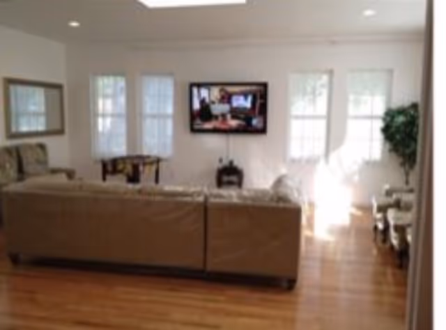Bright living room with a sofa facing a wall-mounted TV, hardwood floors, and windows letting in sunlight.