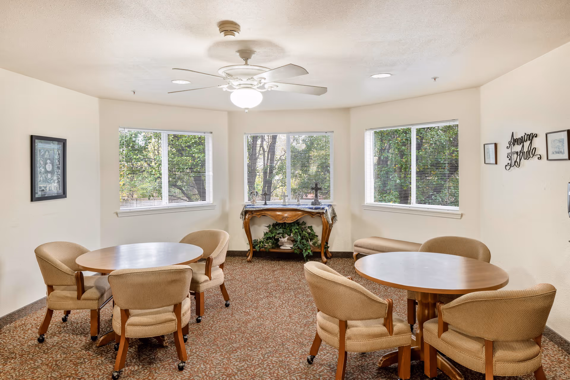 A bright room with three large windows showing green trees outside. Inside, there are two round wooden tables each surrounded by four beige cushioned chairs on wheels. A decorative wooden console table with plants underneath is placed against the wall between the windows. The walls are light-colored with framed artwork and a wall decoration that reads 'Amazing Grace'. The ceiling has a white fan with a light fixture.