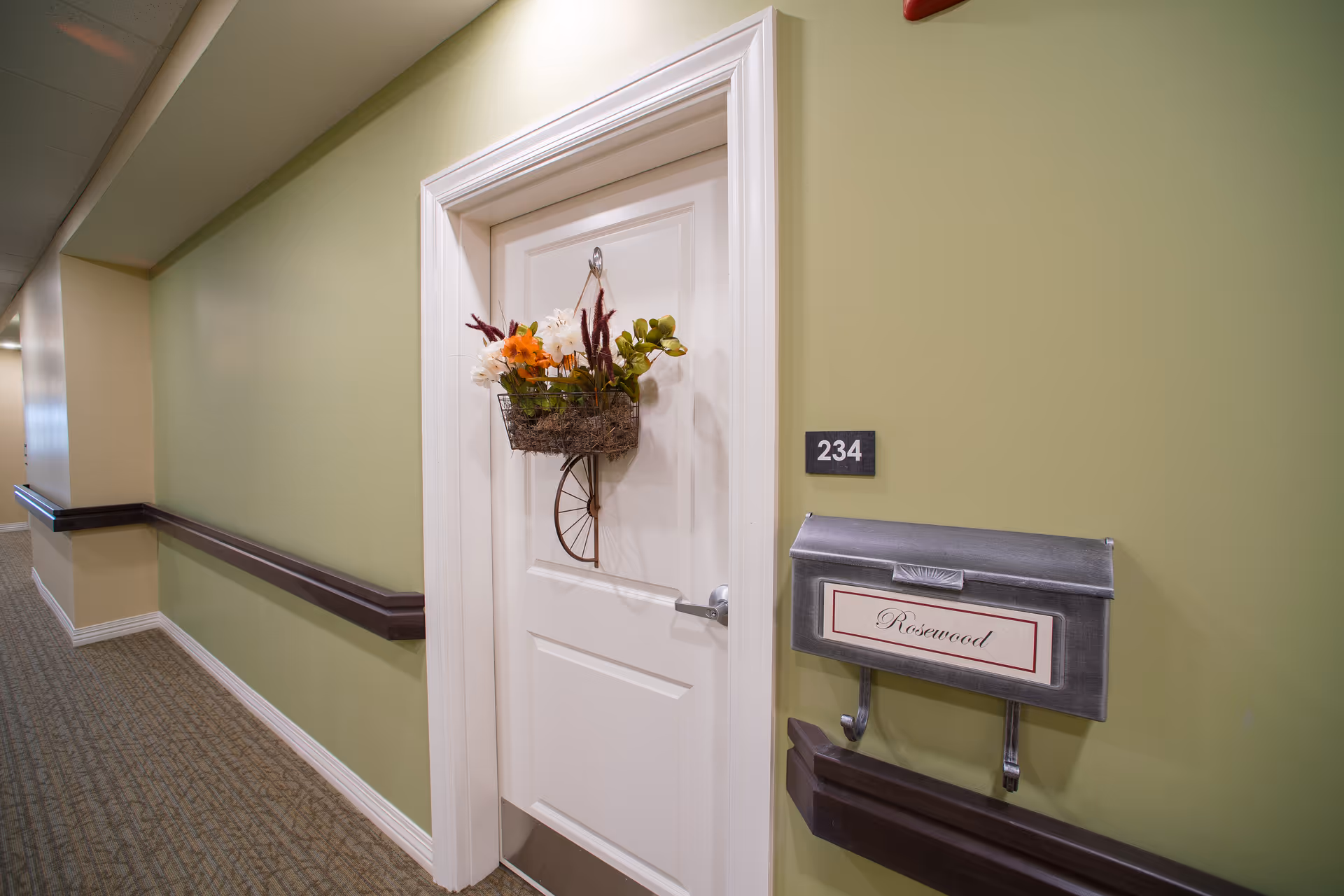 A green-walled facility hallway showing a white door numbered 234 with a hanging flower basket and a mailbox labeled "Rosewood".