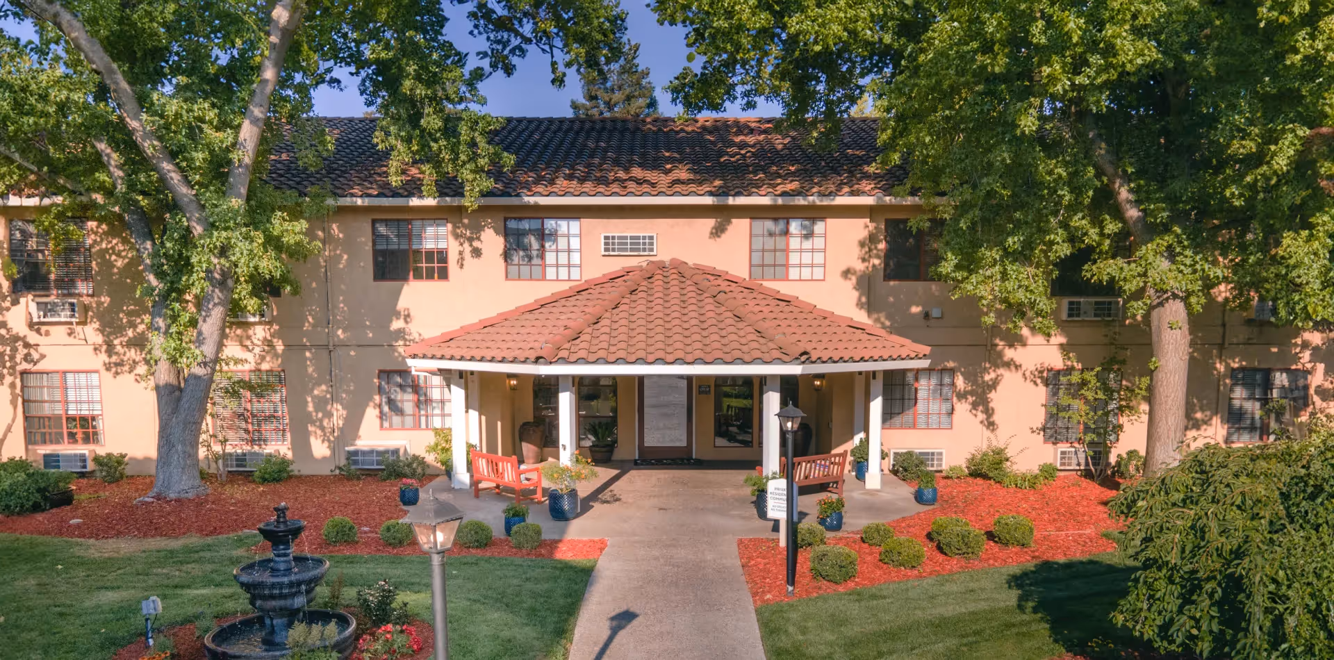 Front entrance of a two-story building with a tiled roof and a covered porch area. The building is surrounded by green trees, manicured bushes, red mulch landscaping, and a small black tiered water fountain in the foreground. There are two red benches on the porch and a lamp post near the walkway leading to the entrance.