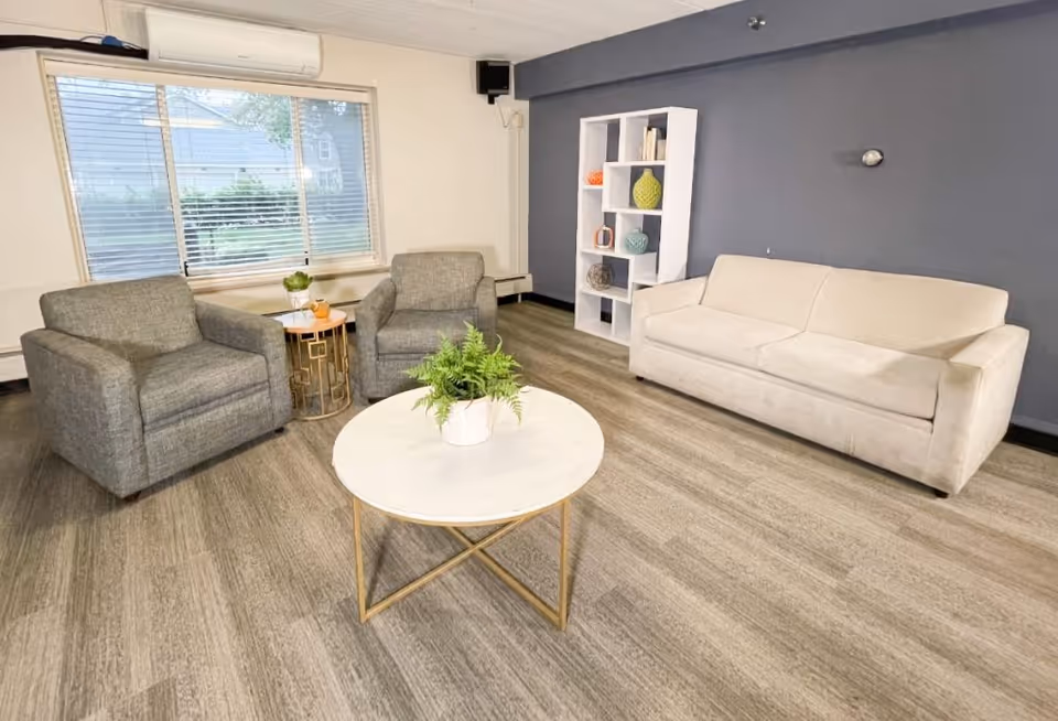 A cozy living room with a beige sofa, two gray armchairs, a round white coffee table with a potted plant, a small side table with a plant and a cup, a white shelving unit with decorative items, and a large window with blinds.