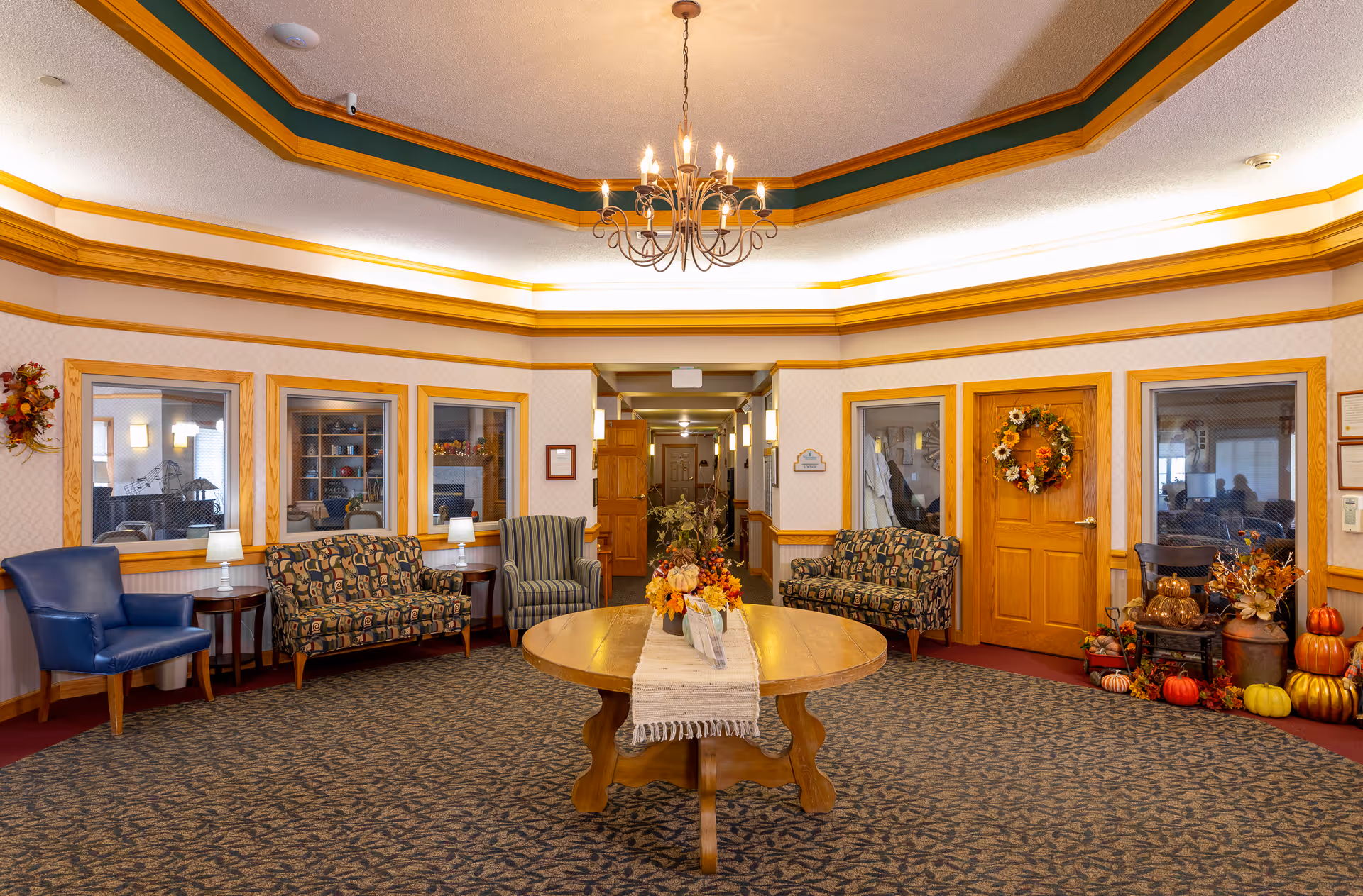 A cozy assisted living facility common area with patterned carpet and a round wooden table in the center decorated with a floral arrangement. The room features multiple armchairs and loveseats with different upholstery patterns, small side tables with lamps, and a chandelier hanging from a tray ceiling with wood trim. The walls have several windows looking into adjacent rooms and a wooden door adorned with a fall-themed wreath. Seasonal decorations including pumpkins and flowers are arranged near the door.