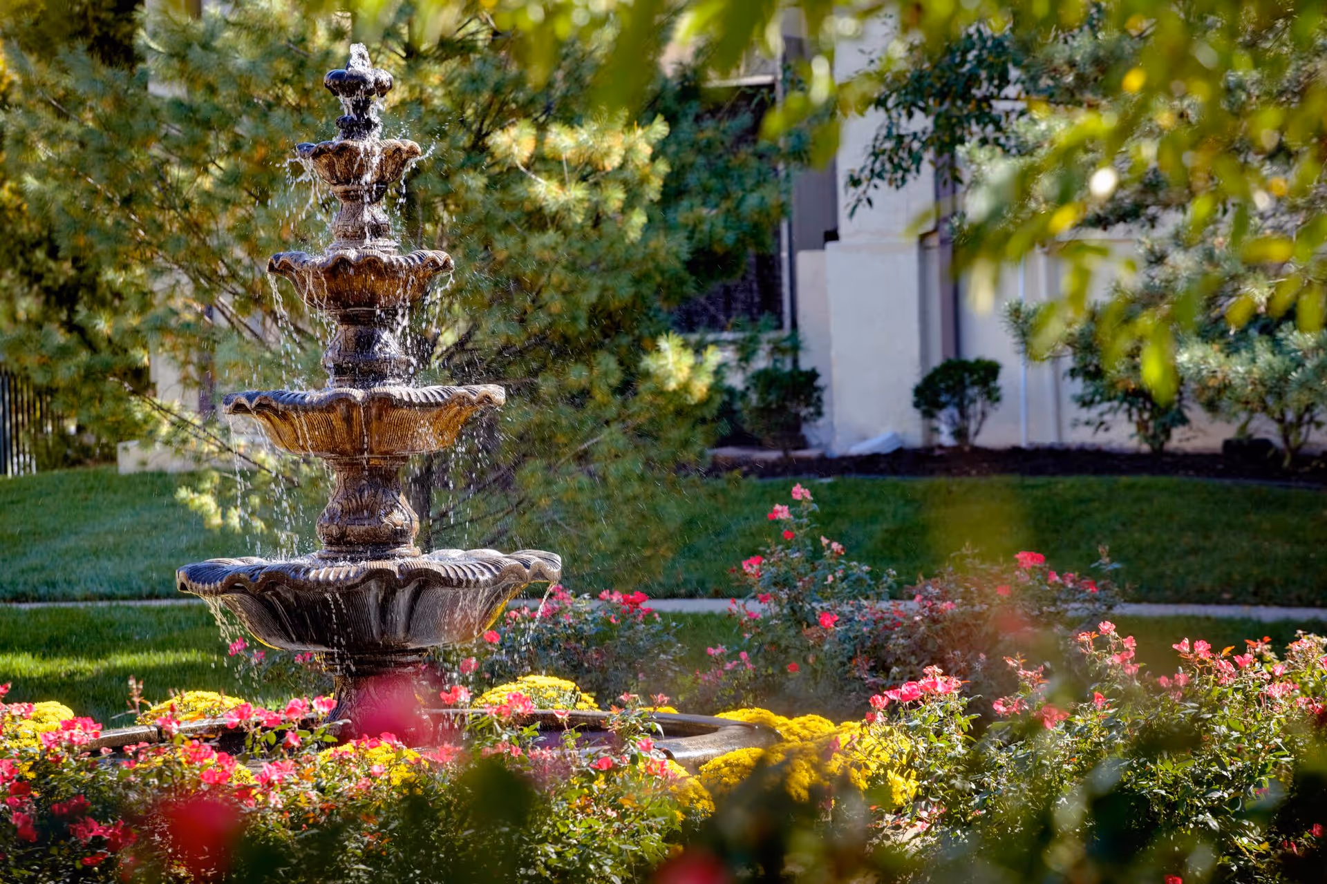 Tiered stone fountain in a landscaped garden with colorful flowers and a building in the background.