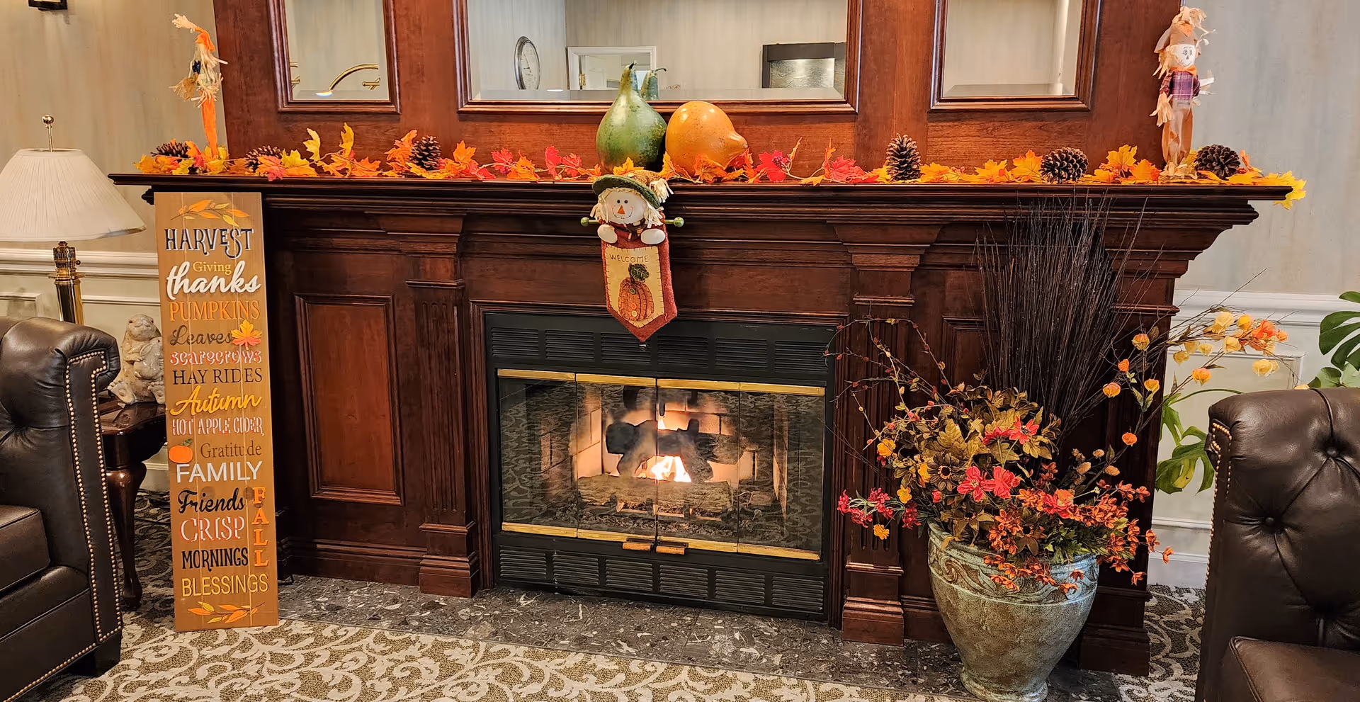 A cozy indoor scene featuring a dark wooden fireplace mantel decorated with autumn-themed items including artificial leaves, pine cones, and gourds. A small scarecrow decoration hangs on the mantel, and a tall fall-themed sign with words like 'Harvest', 'Pumpkins', and 'Family' stands to the left. On the right side, there is a large vase filled with fall foliage and branches. Two dark leather armchairs flank the fireplace, and a table lamp is visible on the left side.