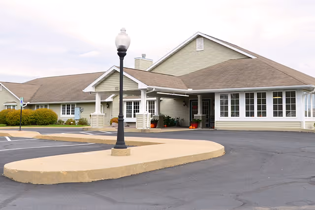 Front entrance of a single-story assisted living building with a covered portico, lamp post, and paved parking area.