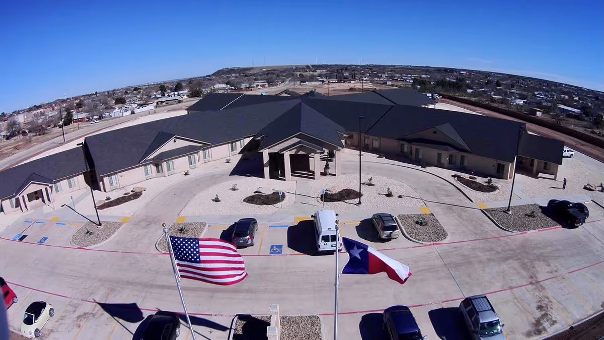 Front exterior of a single-story senior care facility with a circular driveway, parked vehicles, and American and Texas flags flying in front.