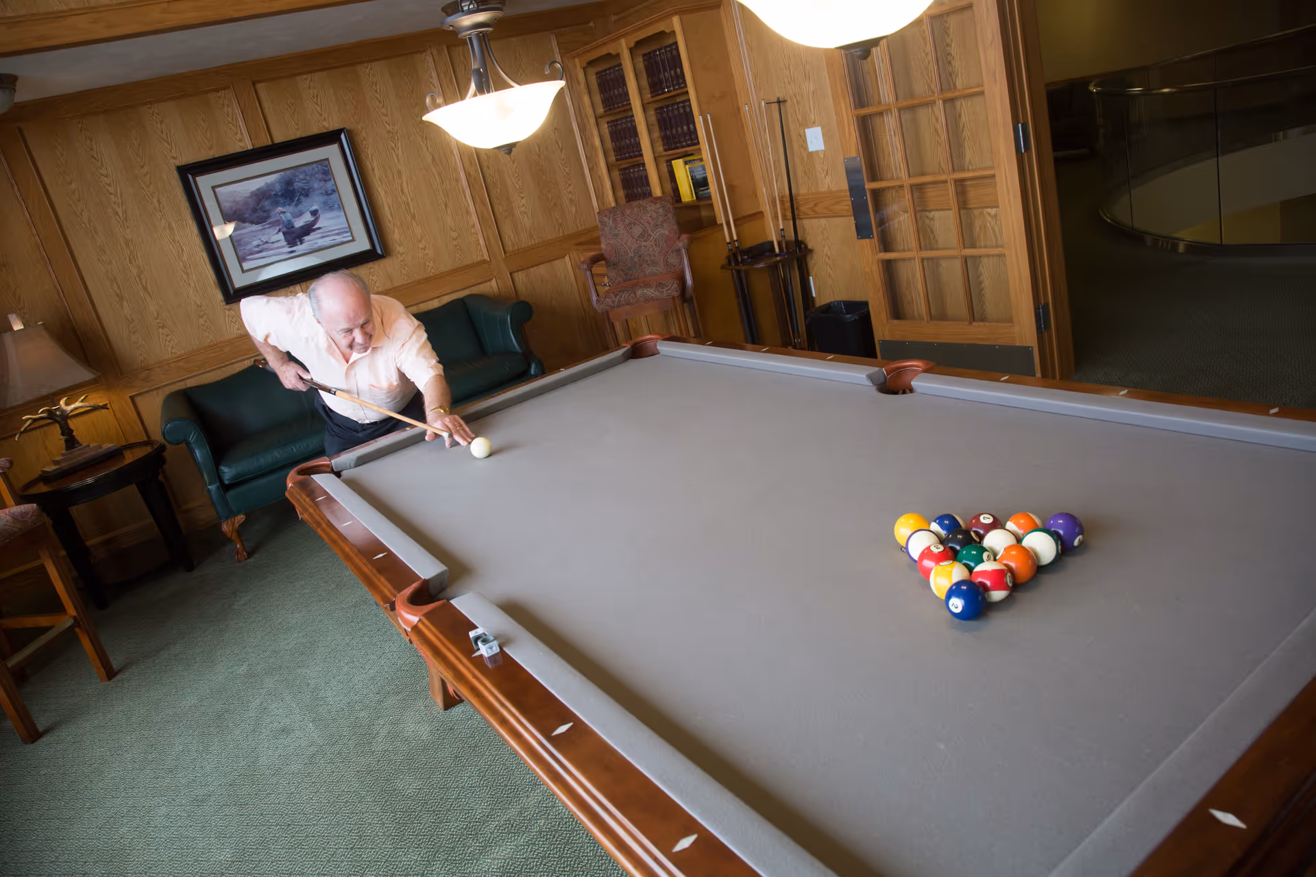 An elderly man playing pool in a cozy room with wood-paneled walls, green carpet, a green leather couch, a framed picture on the wall, and a bookshelf. The pool table has a gray felt surface with a rack of billiard balls set up for a game.