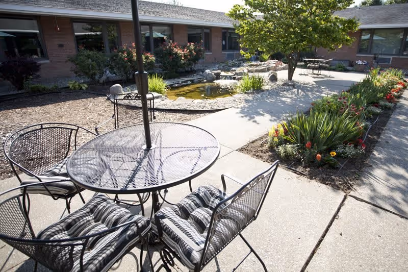 Outdoor patio area with a round metal table and four cushioned chairs. The patio overlooks a small pond surrounded by rocks and greenery, with a tree and flower beds lining the concrete walkway. A single-story brick building with windows is visible in the background.
