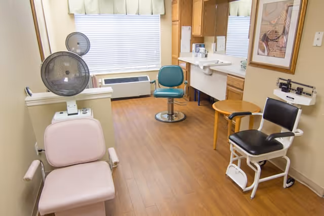 A clinical room with three different chairs including a pink examination chair, a green swivel chair, and a black chair with armrests and a footrest. The room has wood flooring, a large window with blinds and a valance, a sink with cabinets, a small round wooden table, and a framed picture on the wall.