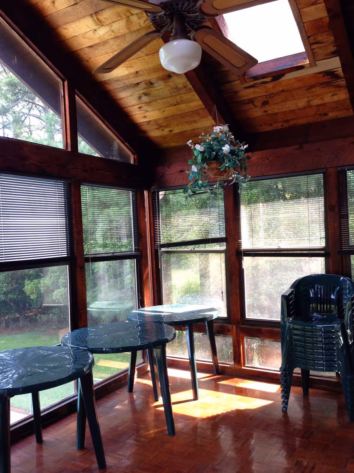 Sunlit room with wooden ceiling and large windows featuring blinds. The room contains three round plastic tables and a stack of green plastic chairs. A ceiling fan with a light fixture and a hanging plant are visible. Outside greenery can be seen through the windows.