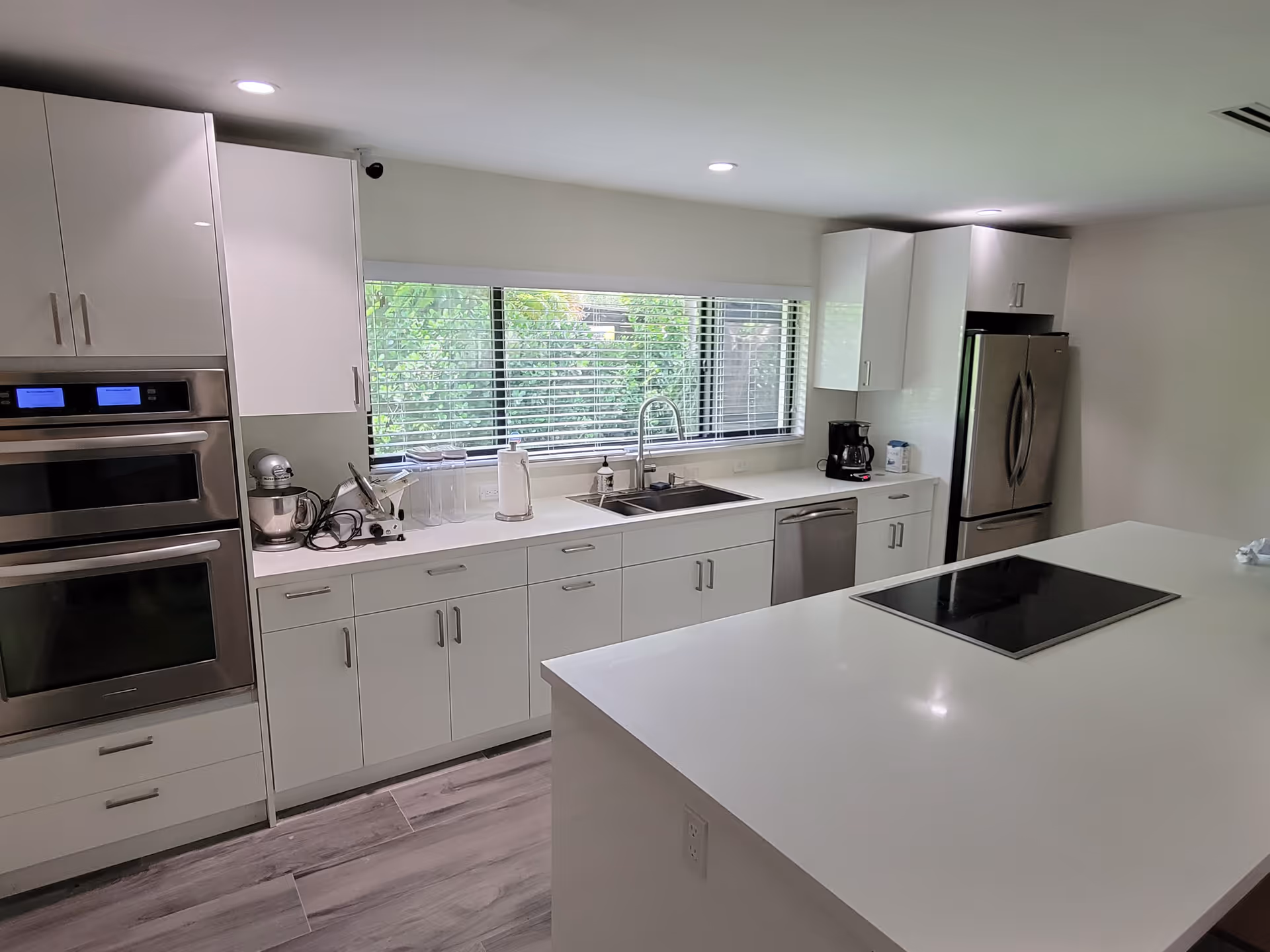 Bright modern white kitchen with a large island, stainless steel appliances, sink under a wide window, and built-in ovens.