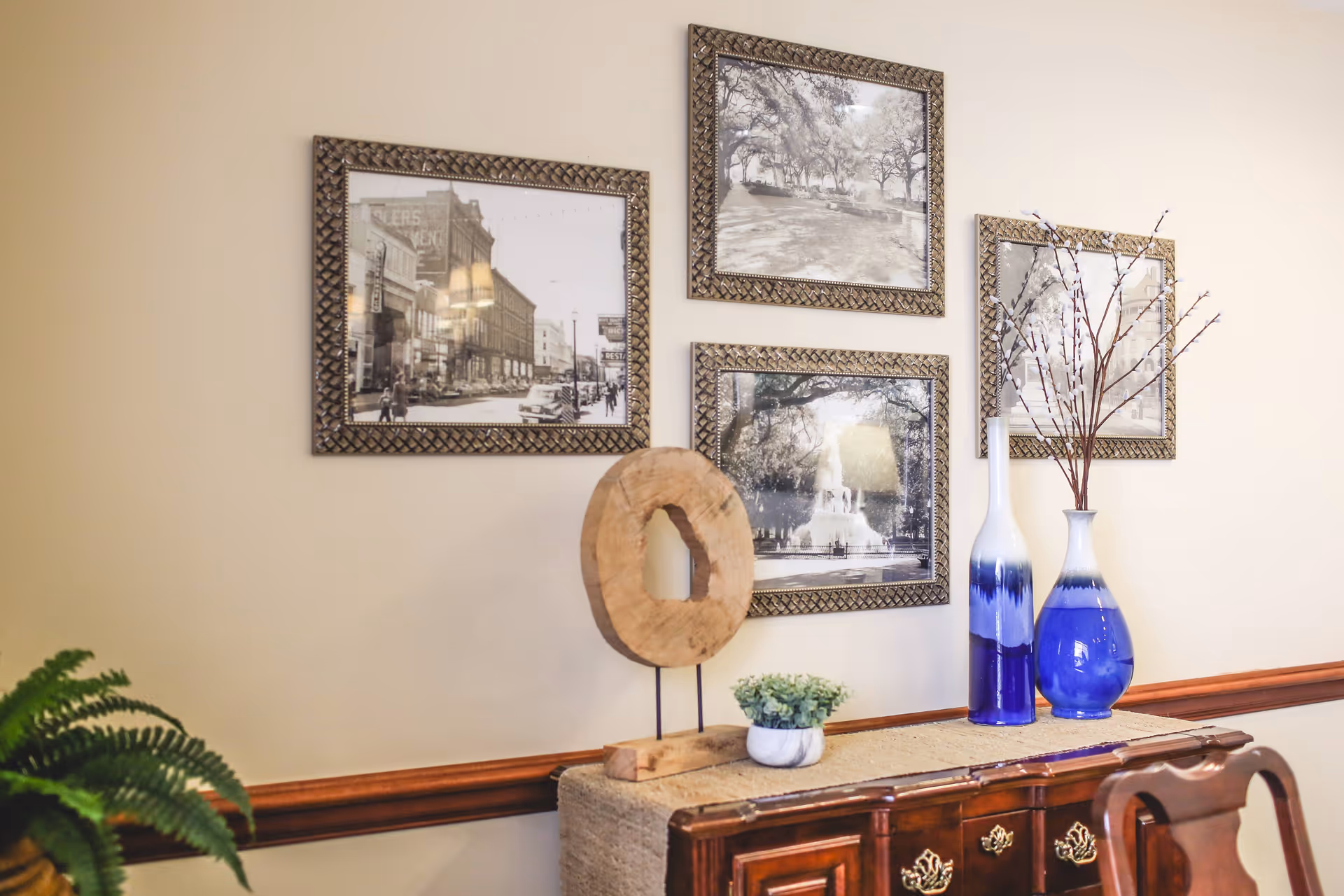 A decorative interior wall featuring four framed black and white photographs arranged in a grid. Below the pictures is a wooden sideboard with a textured runner, a small potted plant, a wooden circular sculpture on a stand, and two blue and white vases with branches. A green fern is partially visible on the left side.