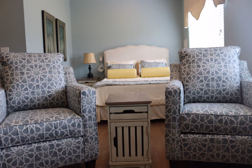 A cozy bedroom with two patterned armchairs facing a bed with yellow accent pillows and a small wooden side table between them.