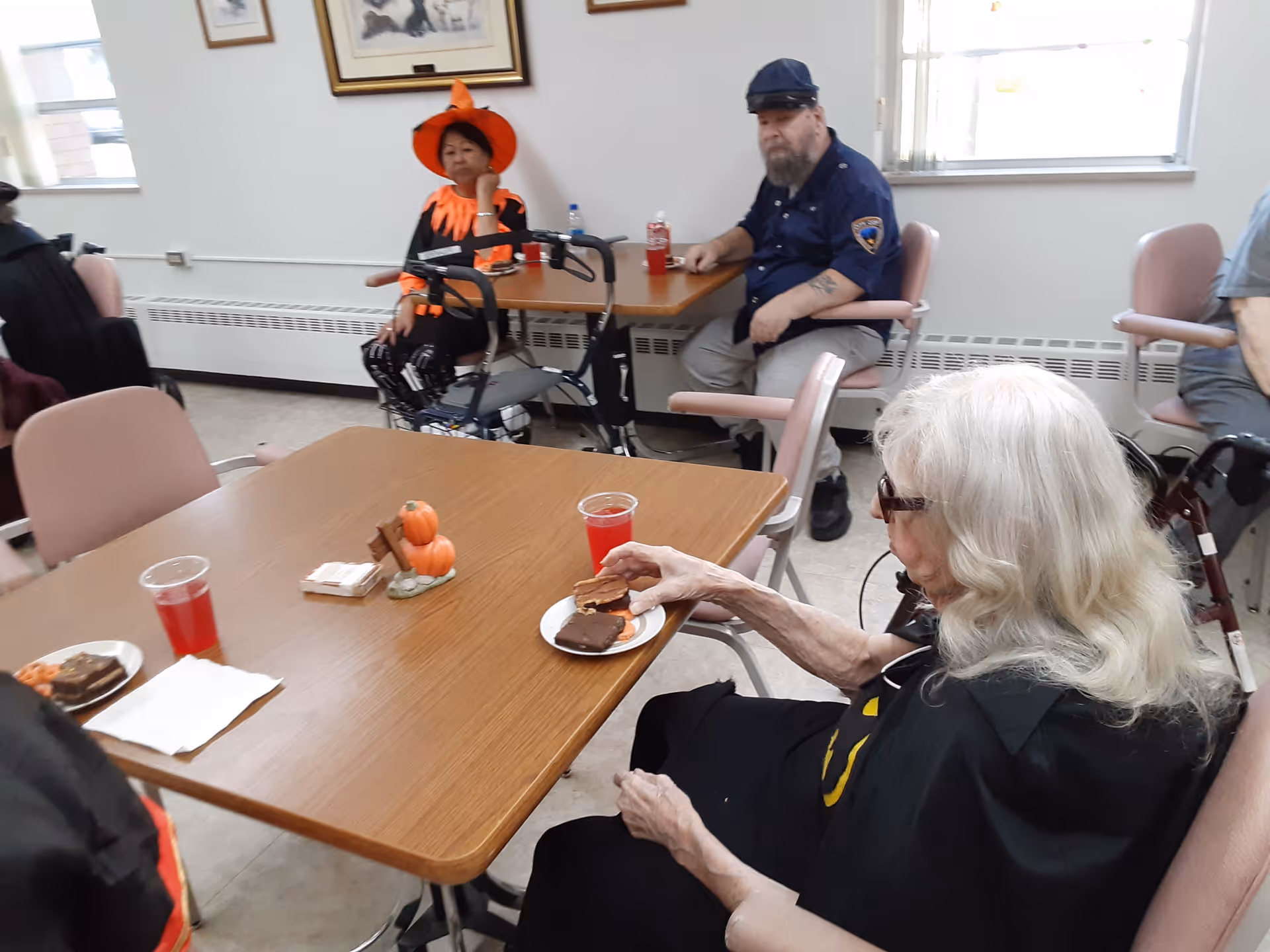 Elderly residents in Halloween costumes seated around tables in a communal dining room enjoying snacks and drinks.