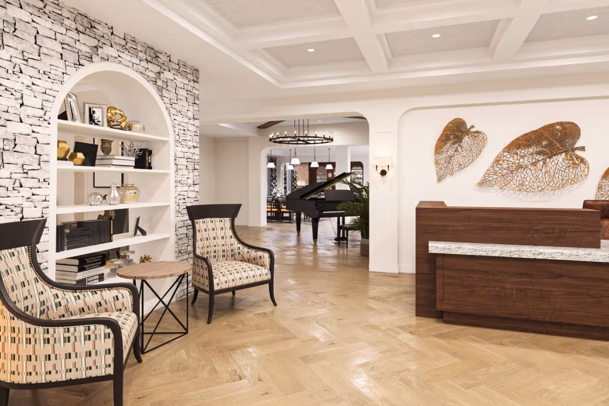 A spacious and elegant interior lounge area featuring two patterned armchairs with dark wooden frames and a small round wooden table between them. To the left is a white built-in bookshelf with decorative items and books. The floor is light wood arranged in a herringbone pattern. In the background, there is a black grand piano and a dining area with pendant lights. On the right side, there is a wooden reception desk with a granite countertop and three large decorative metallic leaf wall art pieces above it. The ceiling has recessed lighting and a coffered design.