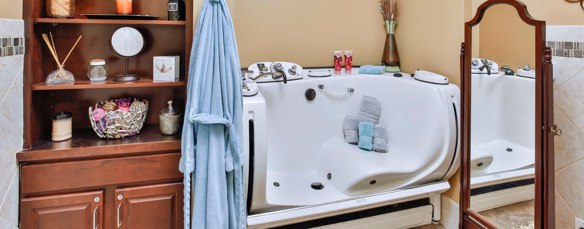 A bathroom featuring a white walk-in bathtub with folded towels placed inside. To the left, there is a wooden cabinet with shelves holding decorative items, a small round mirror, and a blue bathrobe hanging on the side. On the right, a full-length wooden framed mirror reflects part of the bathtub.
