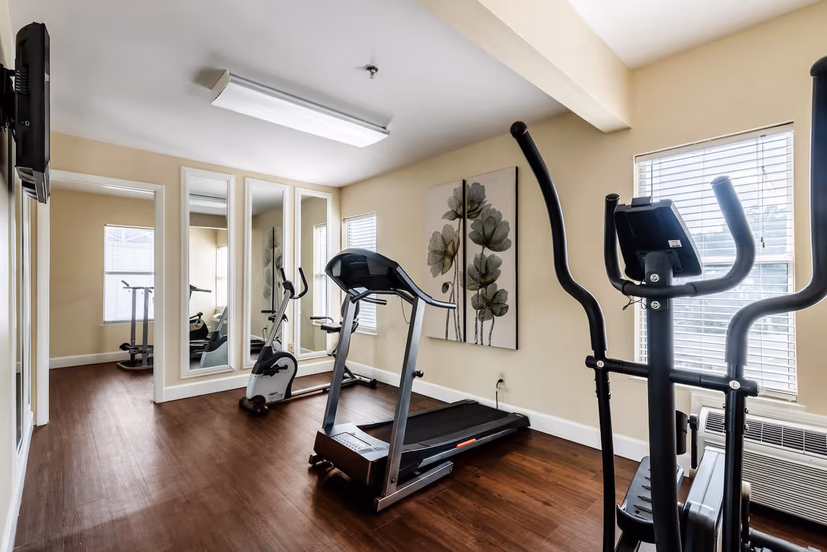 A small fitness room with exercise equipment including a treadmill, stationary bike, and elliptical machine. The room has wooden flooring, beige walls, two windows with blinds, a wall-mounted TV, and a large floral artwork on the wall. There are also three tall mirrors on one wall reflecting the equipment.