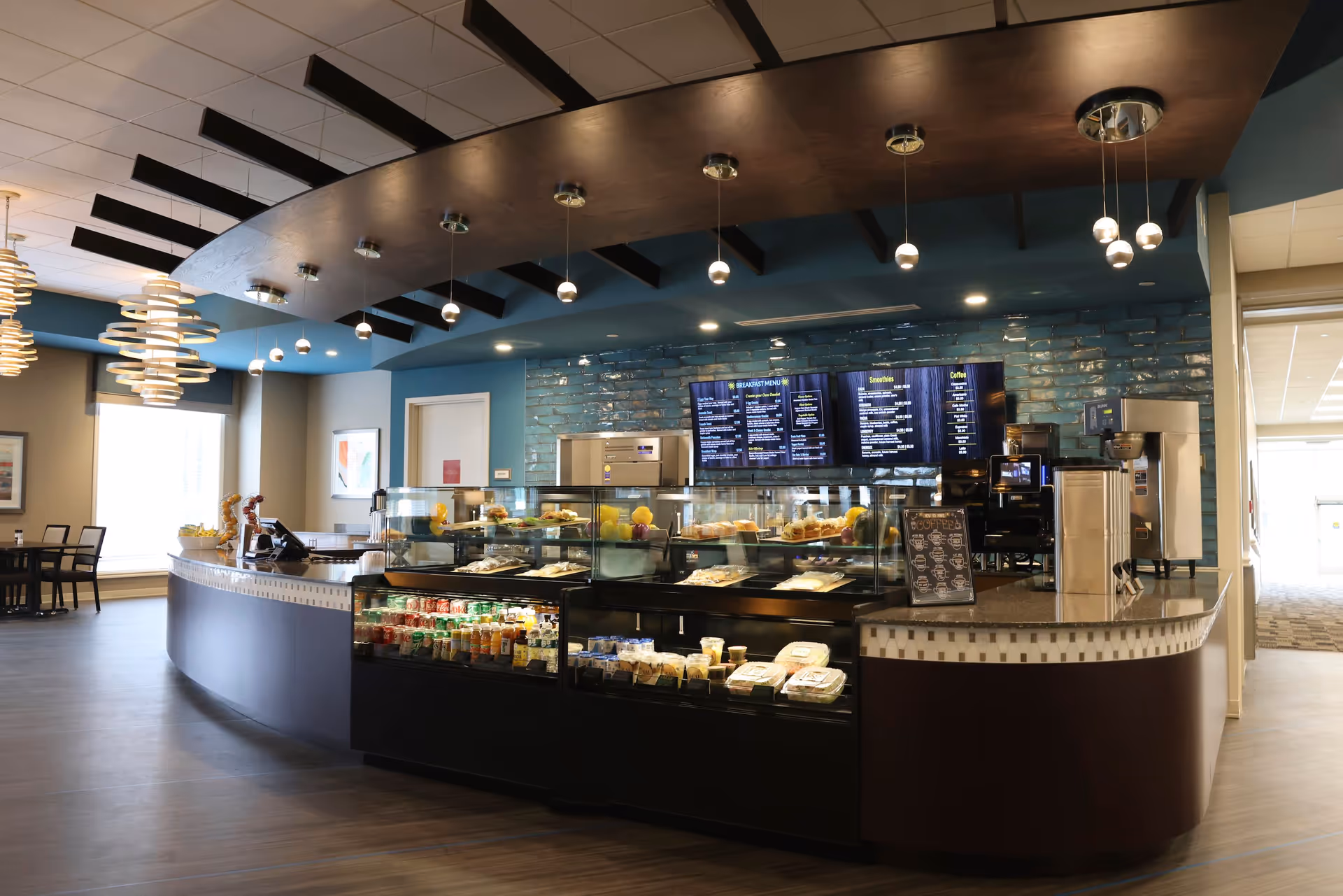 Interior view of a modern cafe or dining area with a curved counter displaying various food items and beverages in glass cases. Above the counter are hanging pendant lights and digital menu boards on a blue tiled wall. There are tables and chairs visible in the background near large windows.