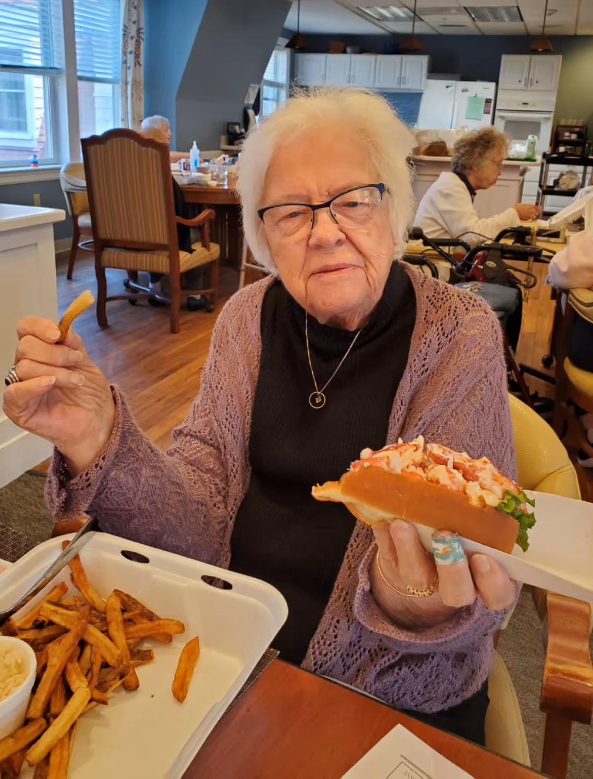 An elderly woman wearing glasses sits at a dining table holding a lobster roll and a french fry with a takeout container of fries in a communal dining area.