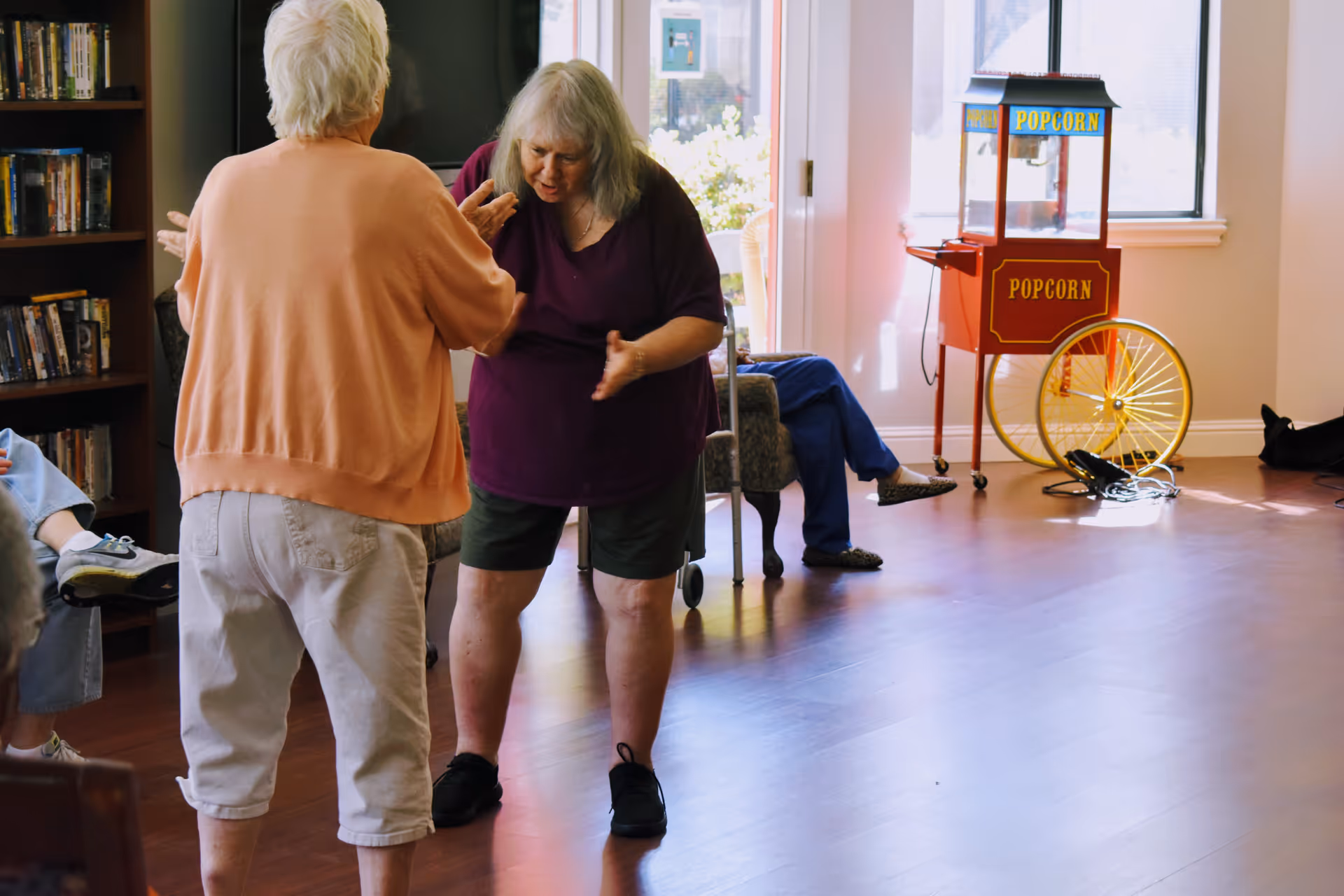 Two elderly women interacting in a common room with wooden flooring. One woman is wearing a peach sweater and white pants, and the other is wearing a purple shirt and dark shorts. In the background, there is a popcorn machine near a window and a seated person partially visible. A bookshelf filled with DVDs or books is on the left side.