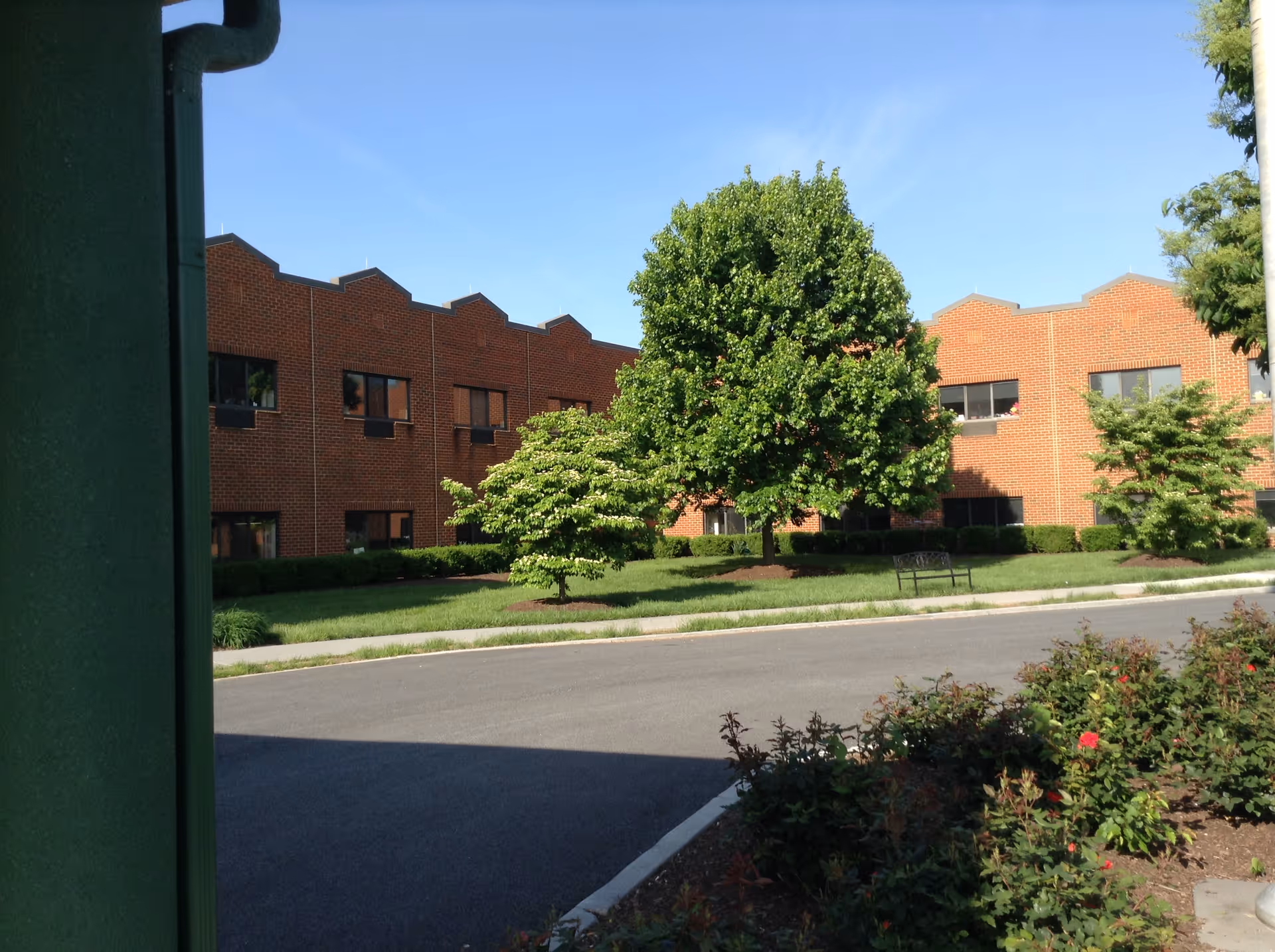 Exterior view of a two-story brick building with multiple windows, surrounded by green trees, bushes, and a well-maintained lawn under a clear blue sky.