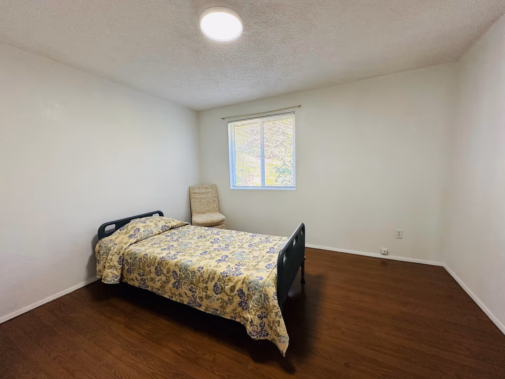 A simple bedroom with a single bed covered in a floral patterned bedspread, a cushioned chair placed near a window with blinds, white walls, a wooden floor, and a ceiling light.