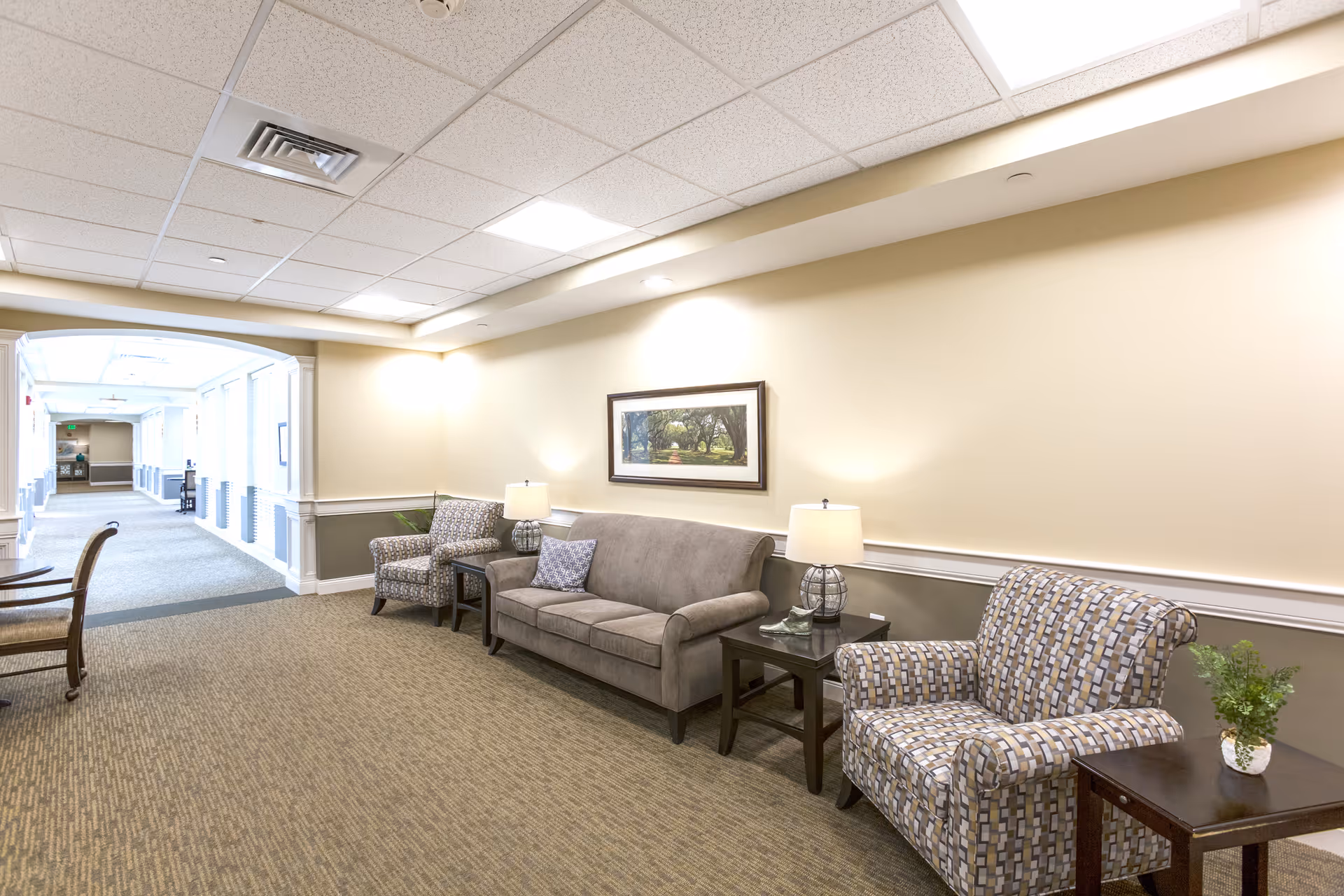 A well-lit interior seating area with a sofa, patterned armchairs, side tables and lamps along a hallway.