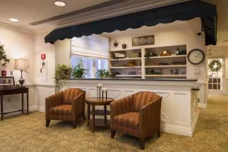 A cozy seating area in a senior living facility featuring two brown upholstered armchairs with a small round wooden table between them, placed in front of a white counter with shelves displaying decorative items. The area is carpeted and well-lit with a lamp on a side table and natural light coming through a window with blinds. A clock and a wreath are visible on the walls.