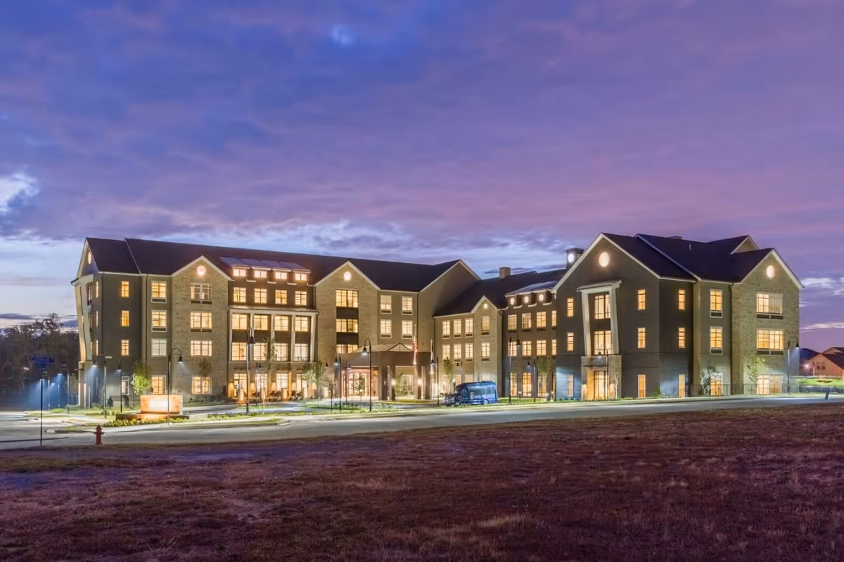 Exterior view of a large, well-lit senior living facility building at dusk with a purple and blue sky in the background. The building has multiple floors with many windows illuminated from inside, and a driveway with a parked vehicle in front.