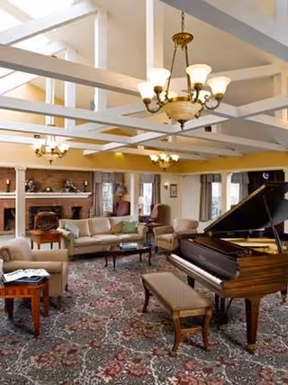 A spacious living room with a grand piano and matching bench in the foreground. The room features a patterned carpet, multiple seating areas with sofas and armchairs, a brick fireplace, and large windows letting in natural light. The ceiling has exposed white beams and several elegant chandeliers.