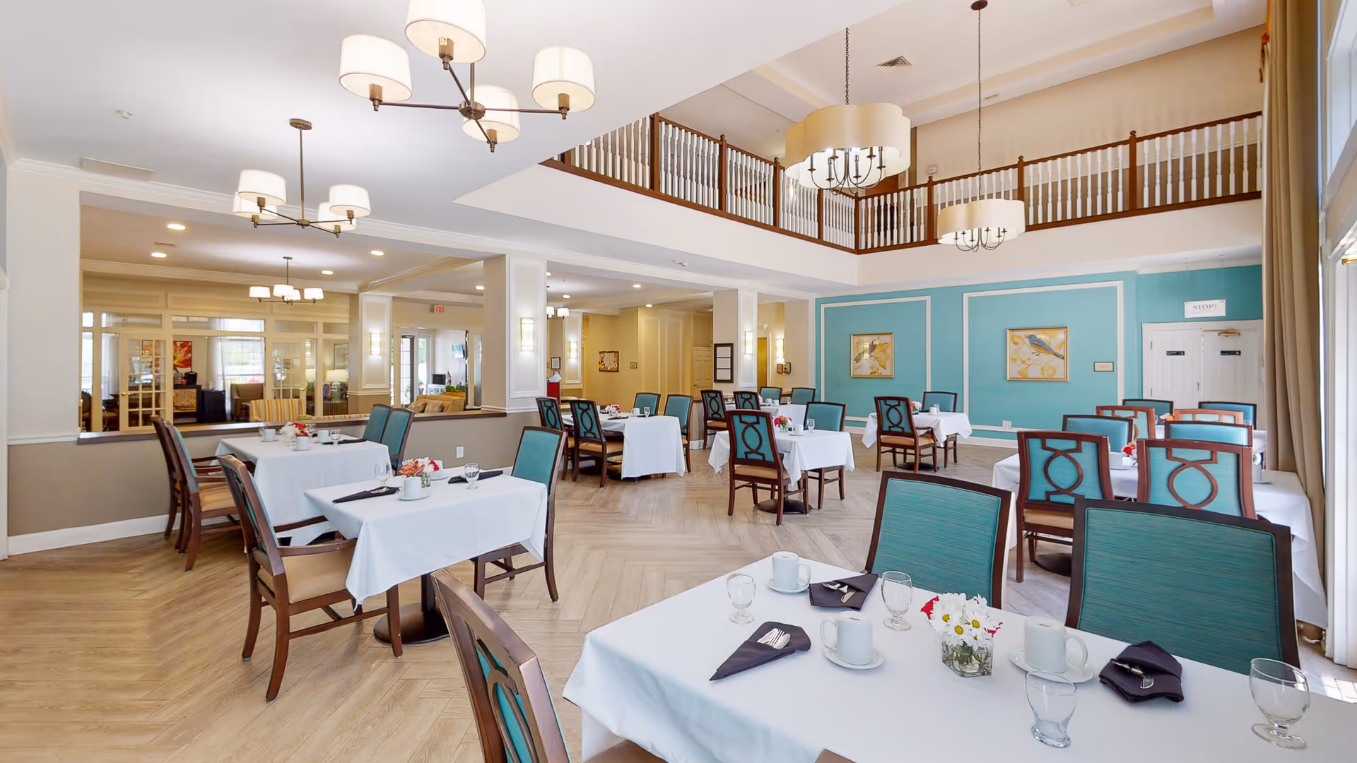 A spacious and well-lit dining room with multiple tables covered in white tablecloths, each set with cups, glasses, napkins, and small flower arrangements. The room features teal accent walls, wooden chairs with teal upholstery, and large windows allowing natural light. Elegant chandeliers hang from the ceiling, and there is a second-floor balcony overlooking the dining area.