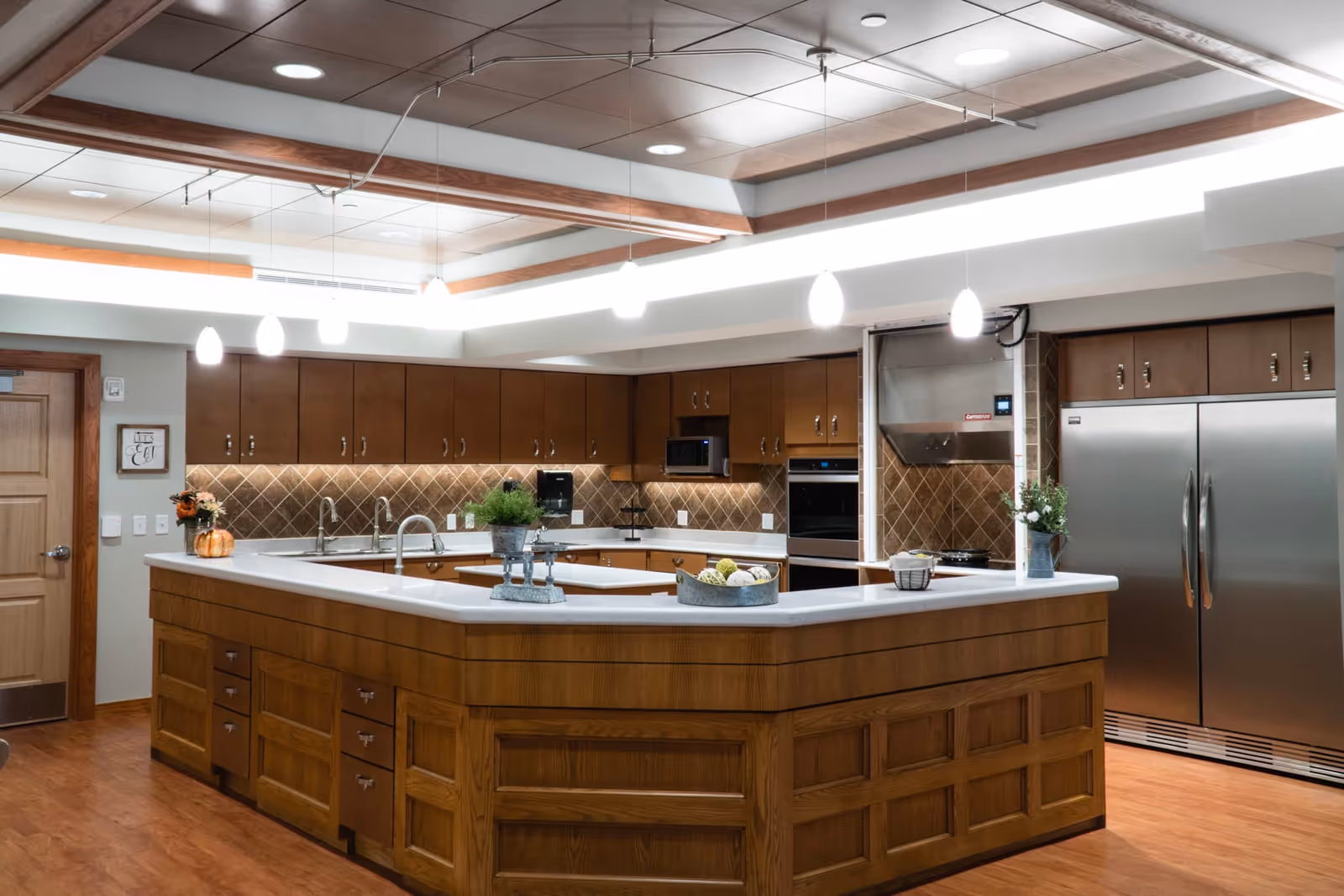 A spacious kitchen with wooden cabinetry and a large white countertop island in the center. The kitchen features stainless steel appliances including a double-door refrigerator, built-in oven, and microwave. Pendant lights hang from the ceiling, illuminating the space. Decorative items such as a small plant, a basket with decorative balls, and a flower vase are placed on the island. The backsplash has a diamond-patterned tile design, and the floor is wooden.