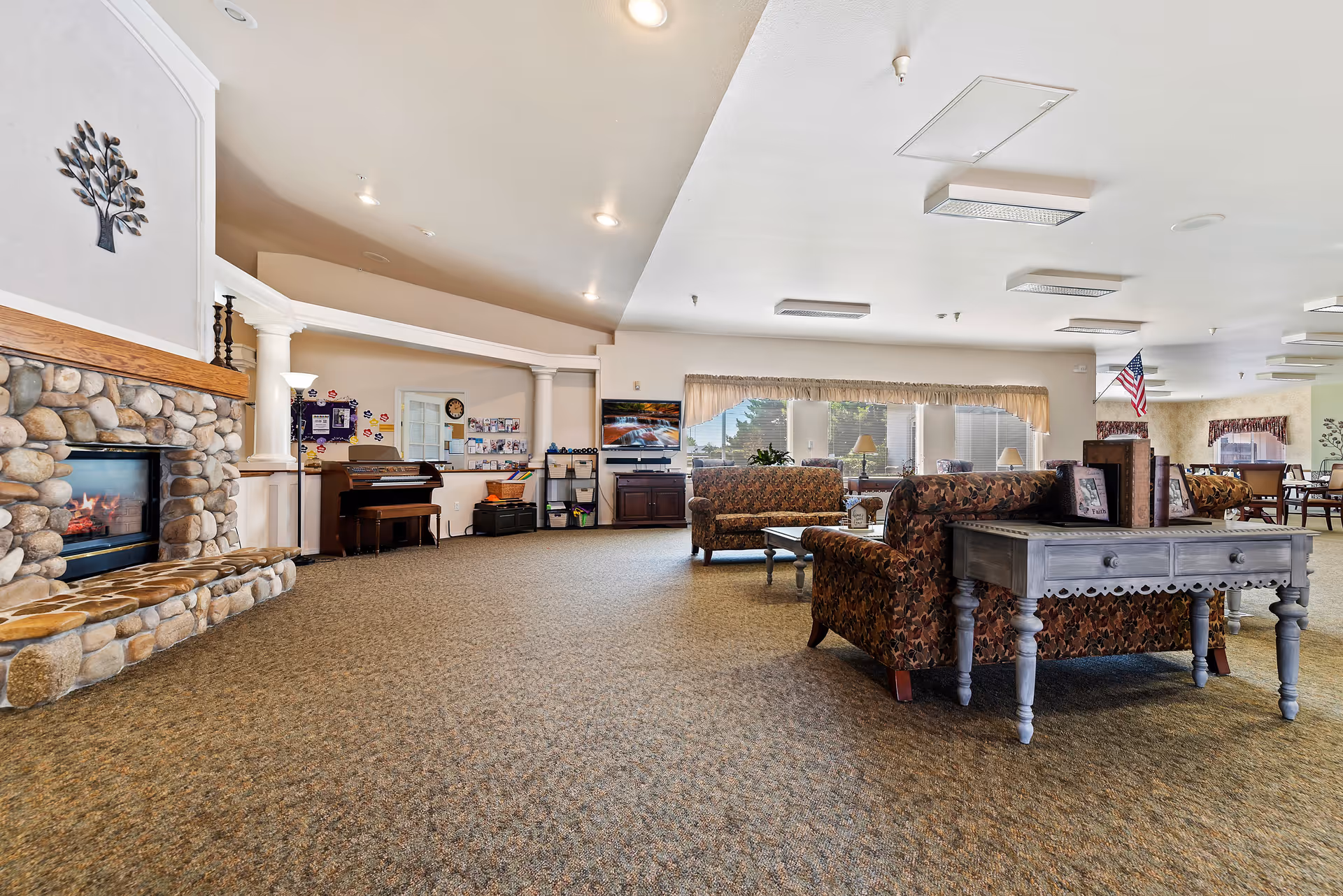 A spacious living room area in Autumn Wind Assisted Living featuring a stone fireplace on the left, patterned upholstered sofas, a wooden coffee table, and a piano against the far wall. Large windows with beige curtains allow natural light to fill the room, and an American flag is visible in the background near a dining area.