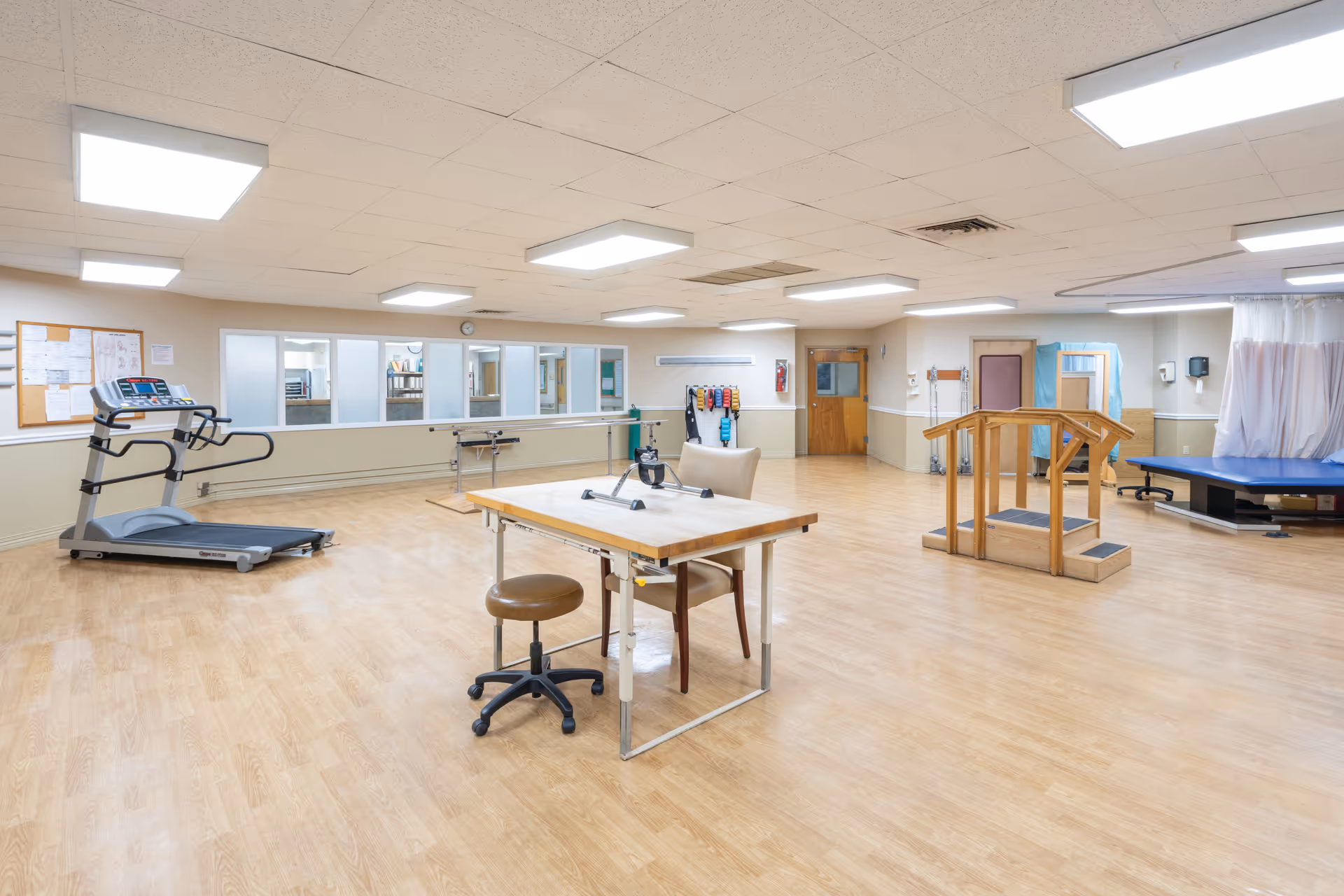 A spacious rehabilitation room with light wood flooring and a drop ceiling with fluorescent lights. The room contains a treadmill, a table with chairs, a small exercise bike, parallel bars for walking practice, a wooden step platform with handrails, and a blue therapy mat with a privacy curtain. There are windows along one wall and a bulletin board with papers pinned on it.