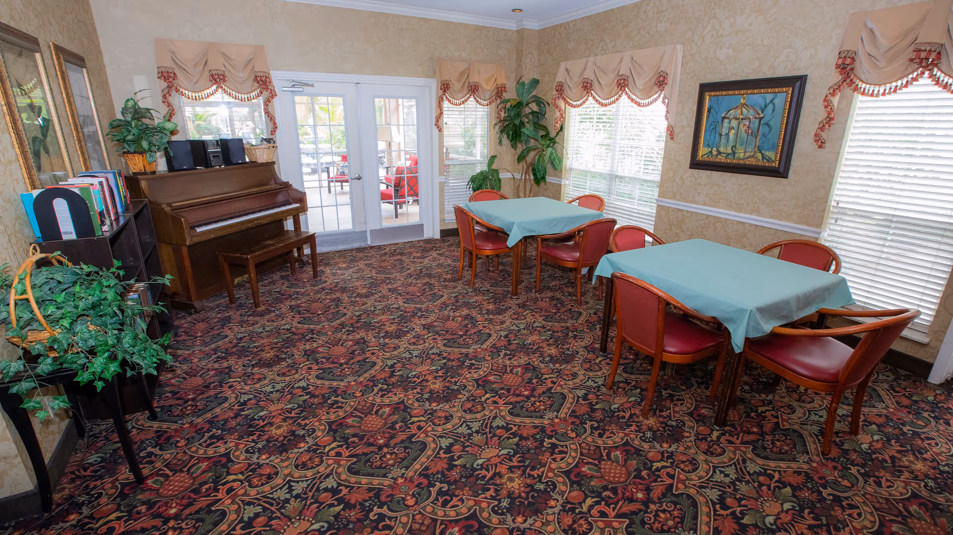 A cozy room with patterned carpet, two tables covered with light blue tablecloths, each surrounded by red cushioned chairs. There is a wooden piano with a bench against the left wall, next to a bookshelf filled with books and a green potted plant. The room has large windows with beige valances trimmed with red tassels, and a framed painting of a birdcage on the right wall. Double glass doors lead to an outdoor patio area with red cushioned chairs.
