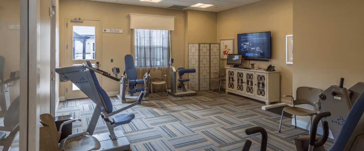 A fitness room in a senior living facility with exercise equipment including a seated leg press machine, a stationary bike, and other workout machines. The room has patterned carpet flooring, beige walls, a window with a curtain, a door, a TV mounted on the wall, and a cabinet with a computer on top.