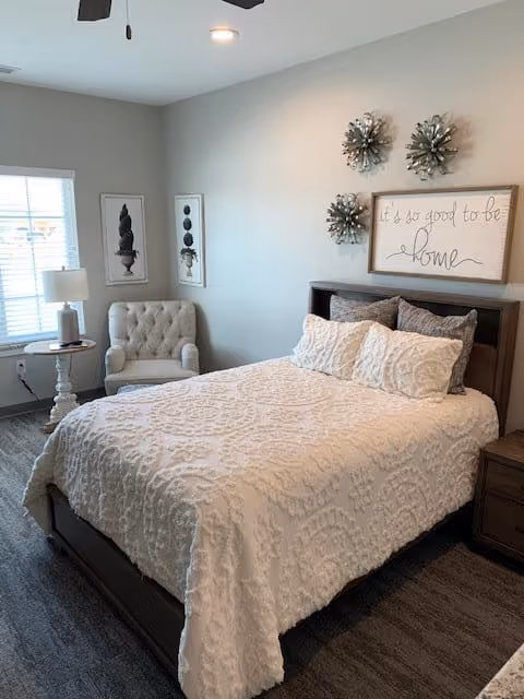 Cozy bedroom with a made bed, decorative pillows, an upholstered armchair by a window, and wall art reading "It's so good to be home."