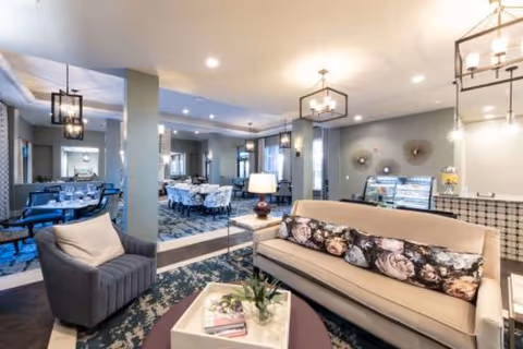 A well-lit interior common area featuring a beige sofa with floral cushions, a gray armchair, a coffee table with a plant and magazines, and a dining area with tables and chairs in the background. The space has modern light fixtures and decorative wall art.