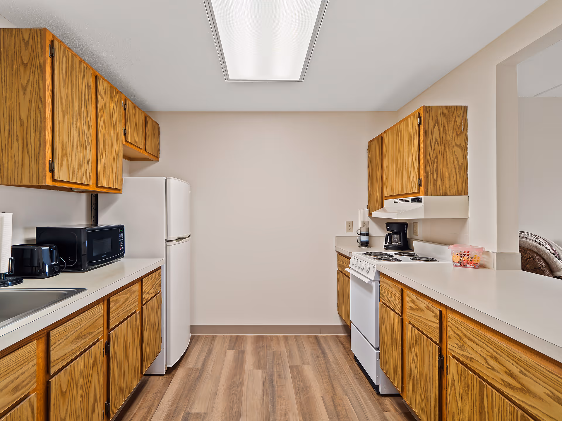 Galley kitchen with oak cabinets, white refrigerator and stove, microwave and coffee maker, and wood-look flooring.